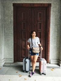 Smiling young woman with luggage standing against house