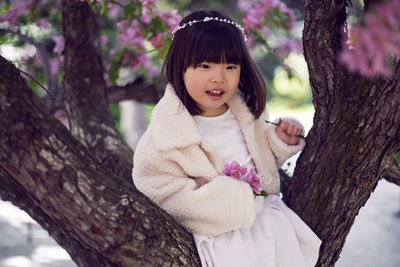 Korean girl in a white light fur coat and a headband sitting on a tree branch in a garden