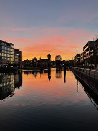 Silhouette buildings by river against sky during sunset