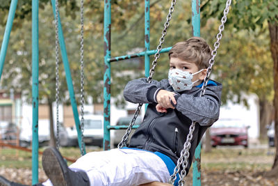 Little boy wearing protective face mask while swinging in the park and looking at camera.
