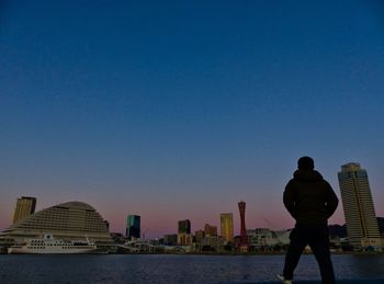 Rear view of man standing by modern buildings against clear blue sky