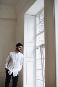 Thoughtful young man with hands in pockets looking through window at home