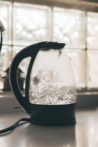 Transparent electric kettle with black handle and lid and with boiling water placed on white table in kitchen