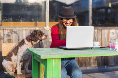 Woman using laptop while sitting on table by dog