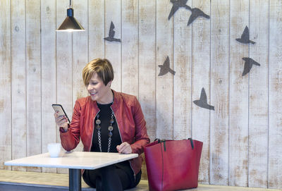 Young woman using phone while standing against wall