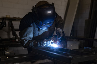 Unrecognizable male employee in protective gloves and helmet using welding machine while working in dark workshop