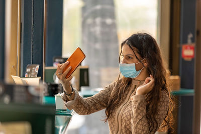 Cute teen wearing medical mask sitting in cafe touching and combing hair using smartphone as mirror