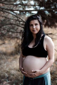 Portrait of smiling pregnant woman with hands on stomach standing in park