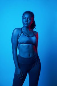 Confident fit black woman in sportswear standing in studio and looking at camera in blue light