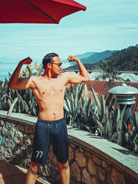 Shirtless man flexing muscles while standing against plants