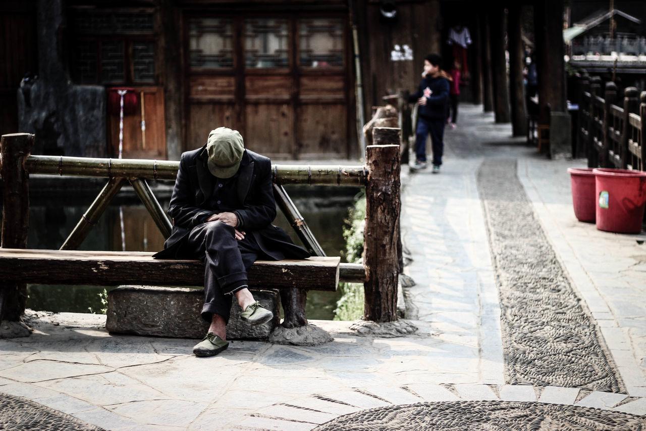 FULL LENGTH REAR VIEW OF MAN SITTING ON BENCH IN CITY