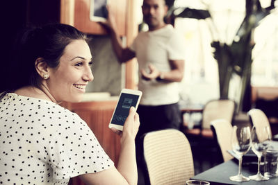 Rear view of smiling female with smart phone looking away while male standing in cafe