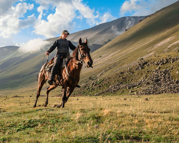 Side view of a shepherd riding on a horse on a mountain valley against sky with clouds