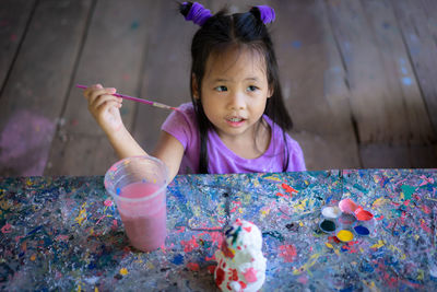 Cute girl holding paintbrush while sitting at messy table