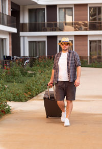 Young caucasian handsome man wearing sunhat, sunglasses with suitcase, coming to hotel.travelling