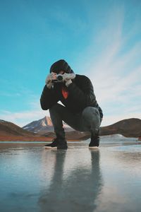 Man photographing through camera while crouching on frozen lake against mountains