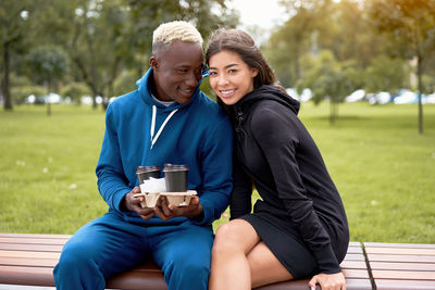 Smiling friends holding coffee cup sitting on bench at park