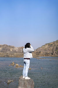 Rear view of man standing on rock by sea against clear sky