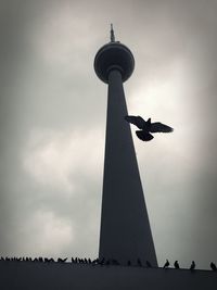 Low angle view of silhouette bird flying by fernsehturm against cloudy sky