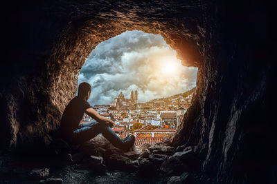 Man sitting in cave while looking at buildings against sky