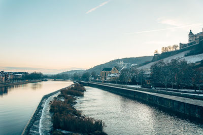 Bridge over river by buildings in city against sky at sunset