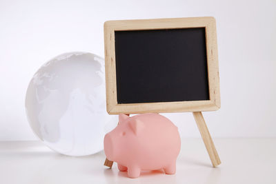Close-up of blank blackboard and globe with piggy bank against white background