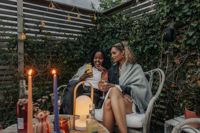 Smiling woman sharing smart phone with female friend sitting on chair in back yard