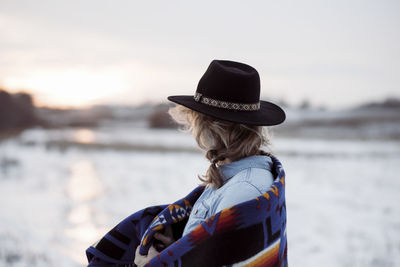 Woman stood in a field looking towards the sunset in a hat & blanket