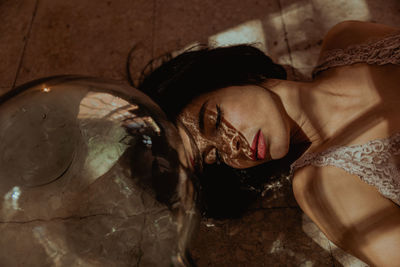 From above of gentle female with closed eyes and in dress lying on floor near glass bottle in room with sunlight
