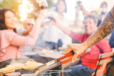 Cropped hand of man preparing food on barbecue grill against people toasting in background