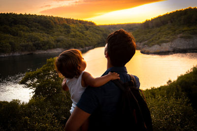 Father holding baby while looking at lake against sky during sunset