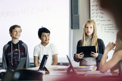 Happy girl giving presentation with digital tablet amidst students in classroom