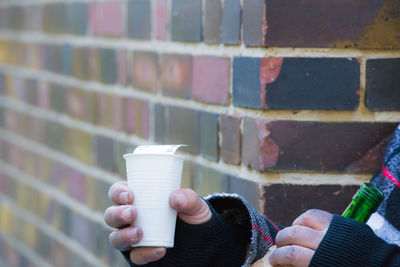 Midsection of man holding paper cup and bottle against brick wall