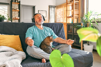 Middle-aged overweight man in wireless headphones relaxing at home with cat and guided meditation.