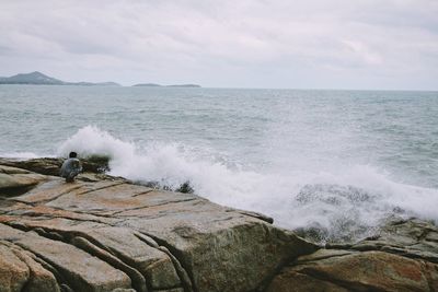 Rear view of man crouching on rock formation by waves rushing against sky