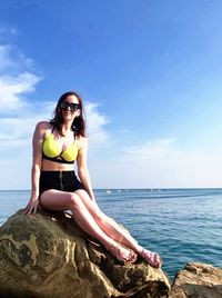 Young woman sitting on rock by sea against sky