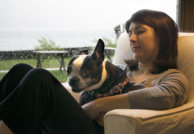 Close-up of woman with dog sitting on chair by wet glass window at home