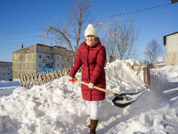 A young woman cleans a path from snow with a shovel near a house on a sunny winter day.