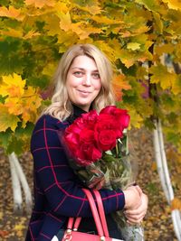 Portrait of smiling woman with red roses standing in park during autumn