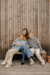 Portrait of smiling woman sitting with sons on bench in front of wooden fence at back yard