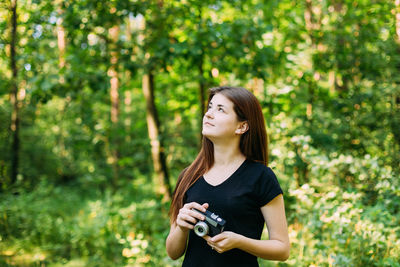 Thoughtful young woman looking up while holding camera against trees