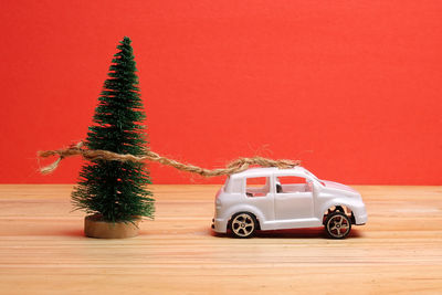 Close-up of small christmas tree tied with toy car on wooden table against red background