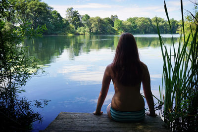 Rear view of topless young woman sitting on jetty against river
