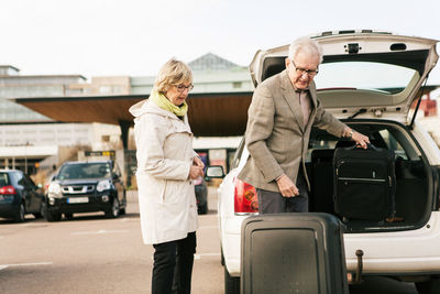 Senior man unloading luggage from car trunk by woman standing in parking lot