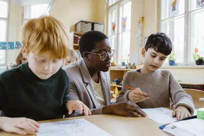 Teacher helping boys in solving problems while sitting at desk in classroom