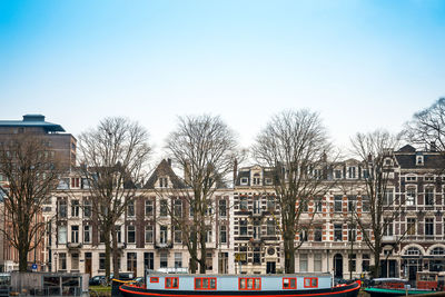 Boats moored by bare trees and buildings against clear sky