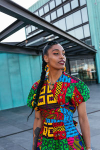 Young ethnic female in braids in colorful clothes looking away while standing near turquoise wall of apartment building in city