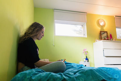 Side view of teenage girl using smart phone while sitting on bed at home