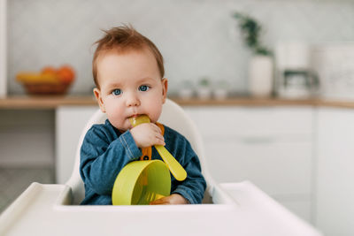 The baby sits in a high children's chair with a spoon and a plate in the kitchen