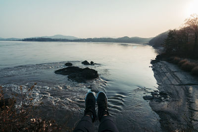 Low section of man standing on rock by lake against sky